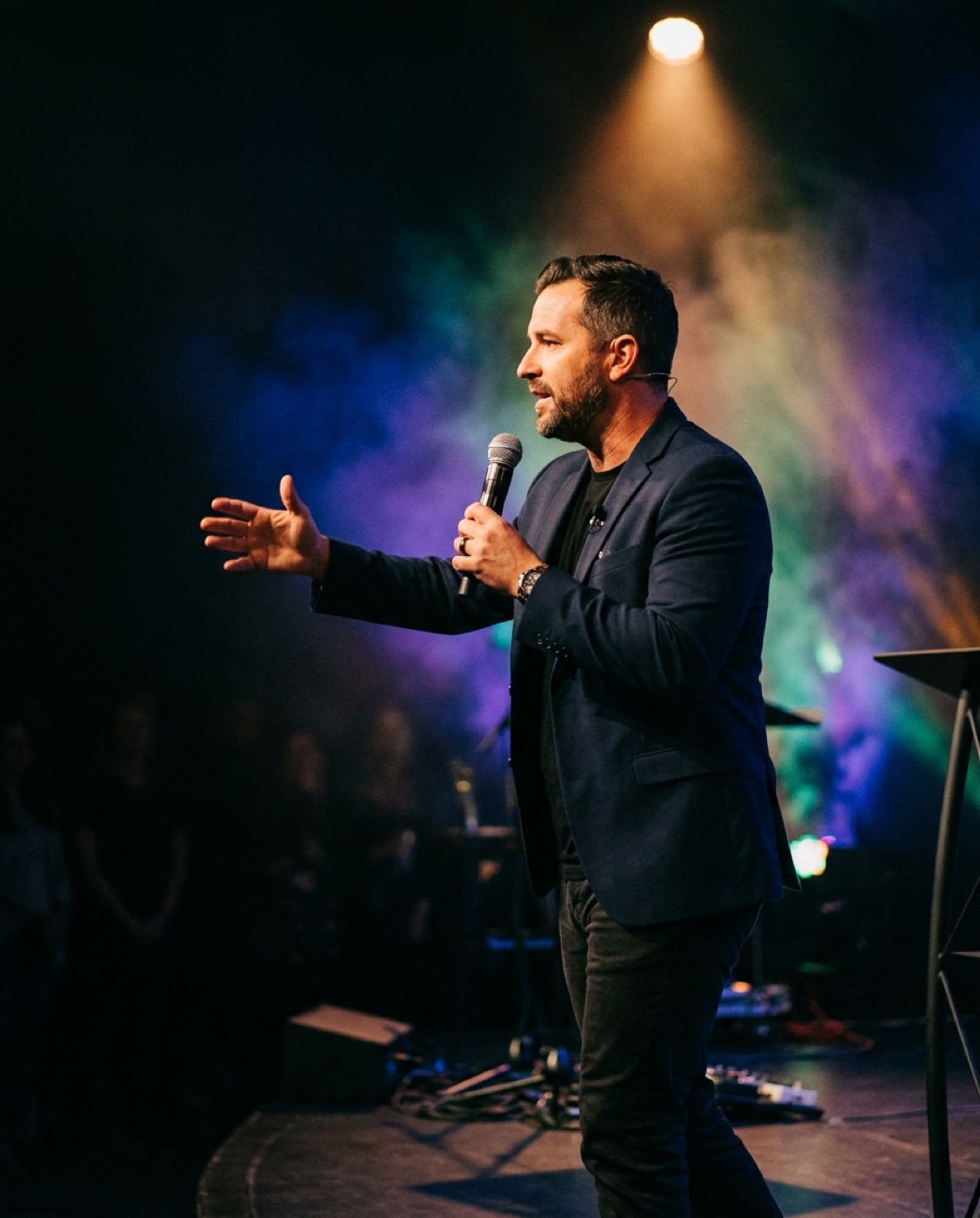A speaker on stage in a navy blazer gesturing with a microphone under coloured stage smoke